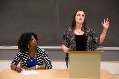 Faculty member standing at podium in front of classroom with another faculty member sitting next to her